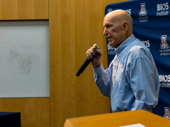 A person is speaking into a microphone at a lectern with the University of Arizona and BIO5 Institute logos displayed in the background.