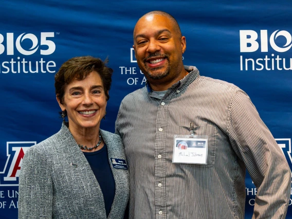 Two individuals smiling at the camera in front of a BIO5 Institute banner at the University of Arizona. One wears a gray blazer, and the other is in a striped shirt with a name tag.