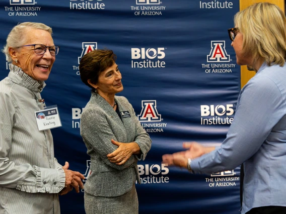 Three individuals are engaging in conversation at the University of Arizona BIO5 Institute, as indicated by the logo-filled backdrop. Two are listening intently while the third, facing away, gestures during the discussion.