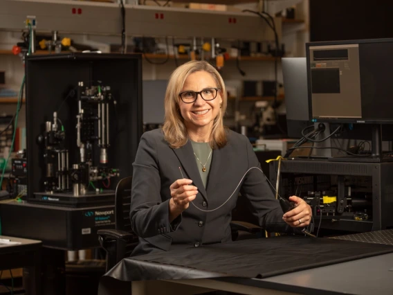 A person in a business suit smiling and sitting at a workbench in a lab with various scientific equipment, holding an optical cable.