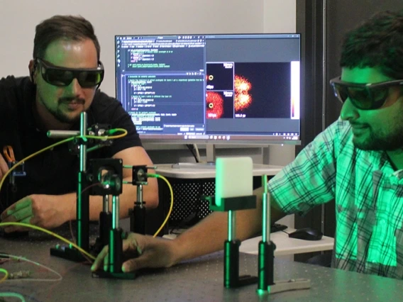 Two individuals in a laboratory setting are working with optical equipment and a computer displaying scientific data. Both are wearing protective glasses.