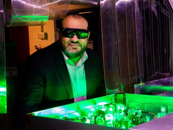 A person wearing sunglasses stands behind a brightly lit green laser system at a laboratory.