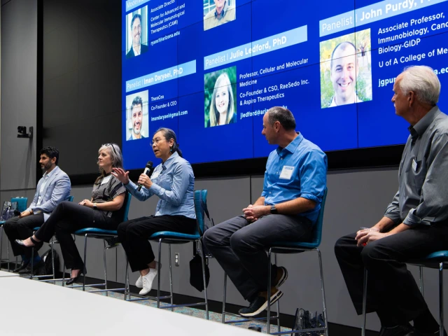 Panel discussion at a conference with six participants seated in front of a digital screen displaying their names and titles. One panelist is speaking while the others listen.