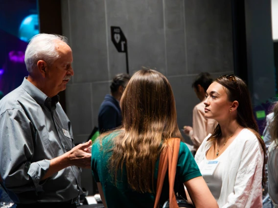 Two people engaging in a conversation at a networking event, with others in the background and ambient lighting.