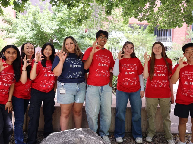 Group of eight students wearing KEYS program t-shirts, smiling and making a 'peace' hand sign outdoors on a sunny day.