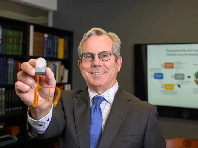 A person in a business suit holding a flexible electronic device in a laboratory setting, with educational presentations visible in the background.