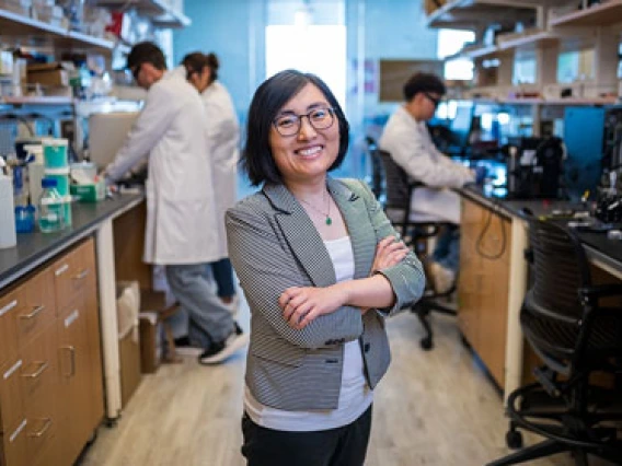 A smiling individual in a gray blazer and green top stands with crossed arms in the foreground of a busy laboratory setting. Several colleagues are working in the background.