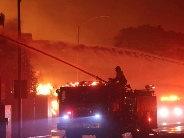 Firefighters on a fire truck using hoses to extinguish a large fire at night, under a smoky, illuminated sky.
