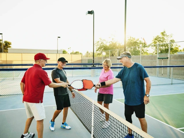 Four adults engaging in a friendly handshake over a net on a pickleball court, with tennis rackets and balls visible, during a sunny day.