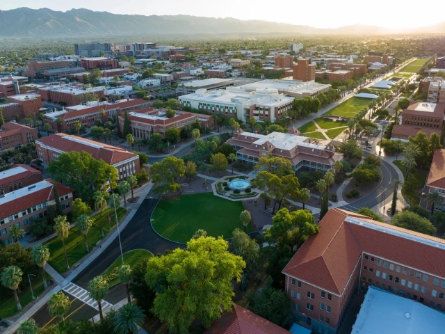 Aerial view of the University of Arizona campus showing red-roofed buildings, green lawns, and surrounding cityscape during sunset.