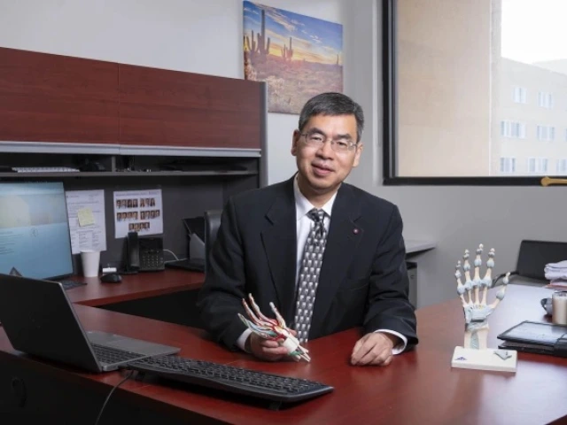 Individual seated at a desk in an office with a laptop, monitors, and various stationery items, smiling towards the camera.
