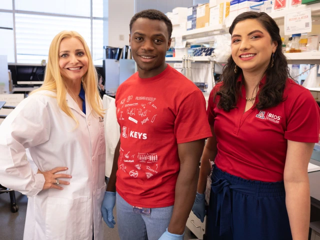 Three people in a laboratory, one wearing a white lab coat and the other two in red shirts, smiling at the camera.