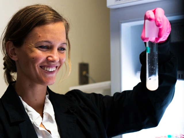 A scientist in a lab coat examines a test tube with a pink substance under a light, smiling with satisfaction.