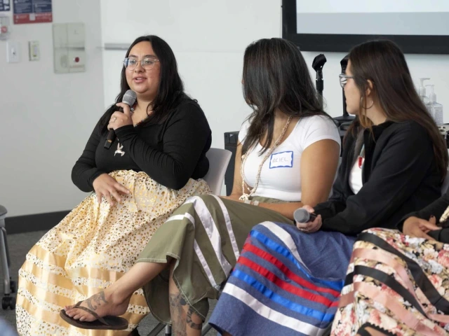 Three individuals sitting on chairs in a panel discussion, with the person in the middle speaking into a microphone. They are in a room with a modern interior design.