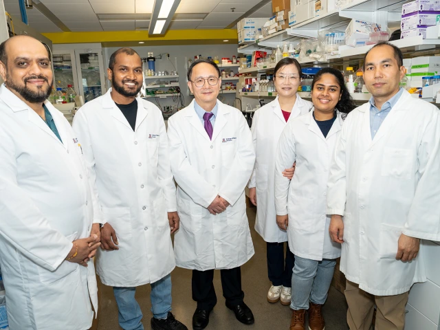 Six researchers in white lab coats standing in a laboratory, with shelves stocked with supplies and equipment in the background.