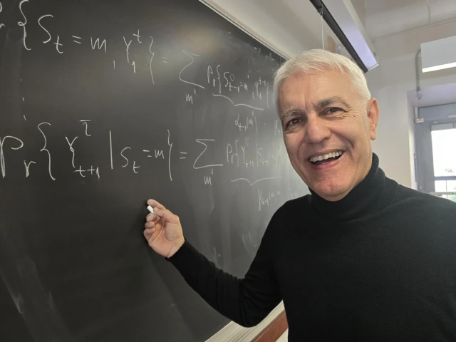 A person smiling while standing in front of a blackboard filled with mathematical equations.