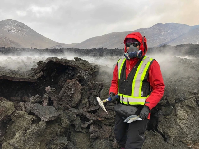 Person in protective red gear, including a gas mask and helmet, holding a tool, stands near a smoking lava field in a volcanic landscape.
