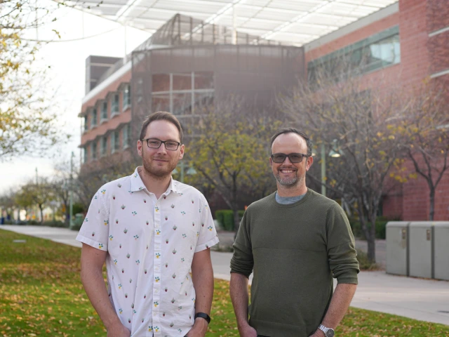 Two people stand smiling on a pathway, with a brick building and trees in the background.