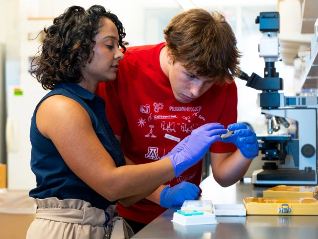 Two individuals are in a laboratory setting with one wearing a red shirt with various logos, including the University of Arizona logo, and both focus on a slide. 