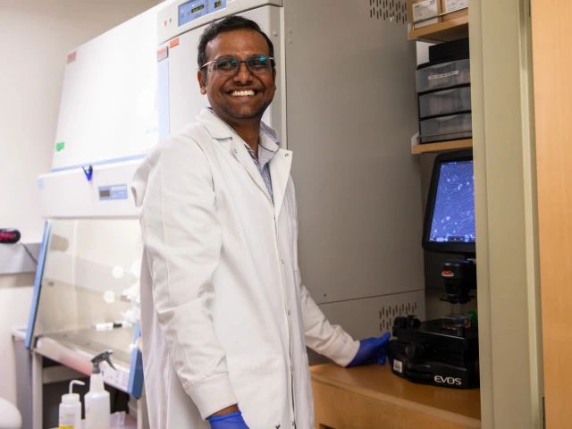 Person in a lab coat and gloves smiles while standing beside a microscope in a laboratory setting.