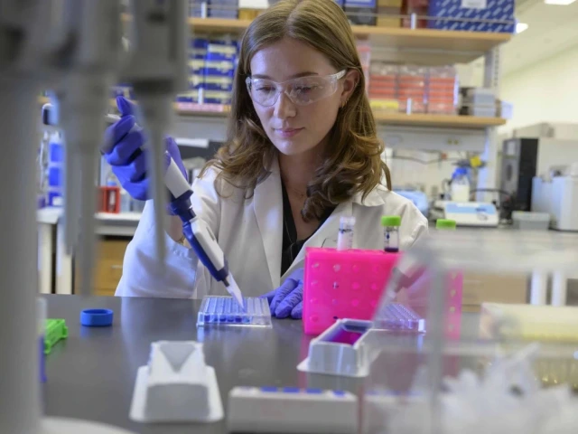 A person in a lab coat and safety goggles is using a pipette in a laboratory setting. The background features shelves with various lab supplies and equipment.