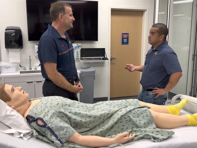 Two individuals in a medical training room are discussing near a hospital bed with a medical training mannequin lying on it. The room contains medical equipment and monitors.