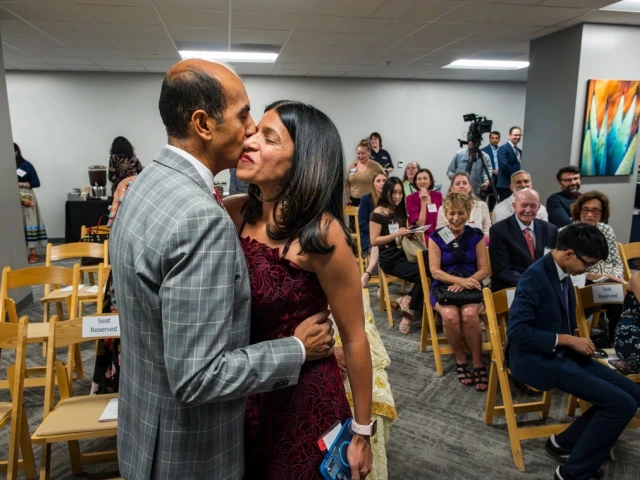 A person in a gray plaid suit and another in a red patterned outfit are embracing in a room filled with seated people. Some seats have "Seat Reserved" signs. In the background, attendees are seated, holding programs and conversing. A colorful abstract painting is mounted on the wall, and there is a camera on a tripod.