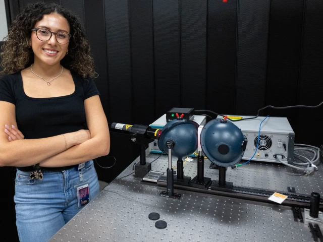 A person with curly hair and glasses stands smiling next to a laboratory setup involving lasers and optical equipment. 