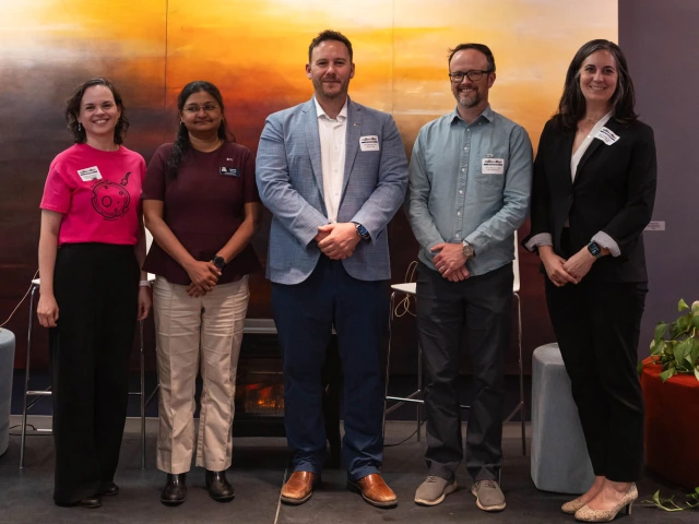 Five people stand in a row, smiling, in front of a large abstract mural, dressed in business casual attire and each has a name tag.