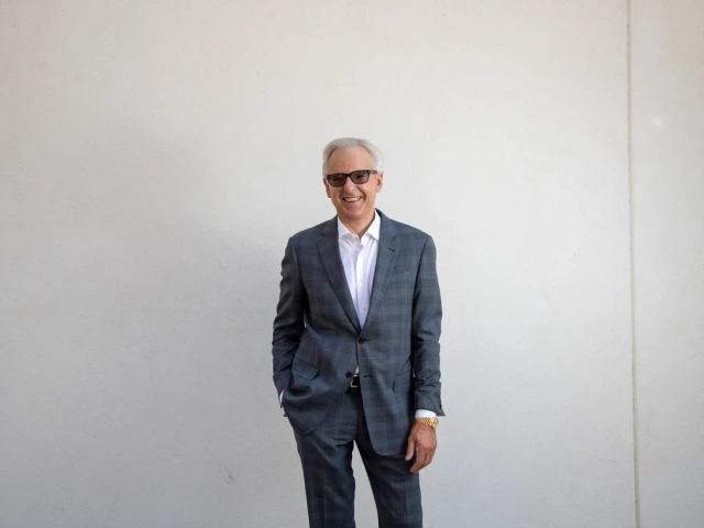 A person standing against a plain white wall, wearing a plaid suit, glasses, and a smile.