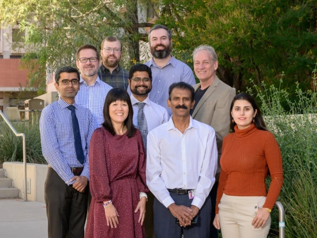 A group of nine people standing outdoors in front of a building and green foliage. They are dressed in business casual attire, and smiling at the camera.