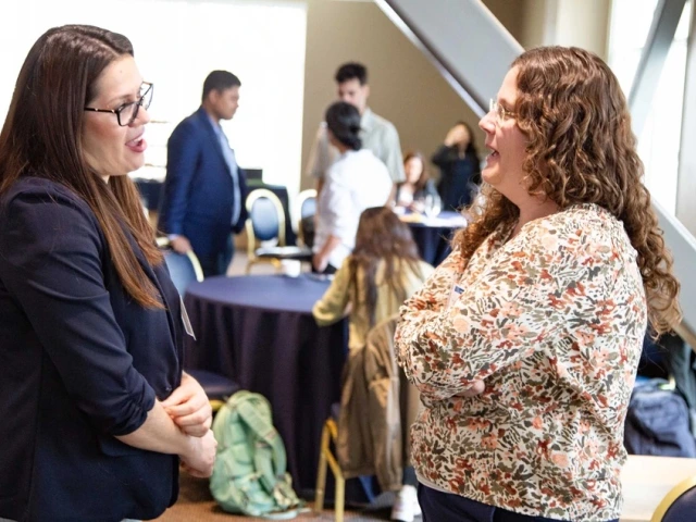 Two people are engaged in conversation at an indoor event. They stand near a table with chairs, and several other people are interacting in the background. The setting appears to be professional.