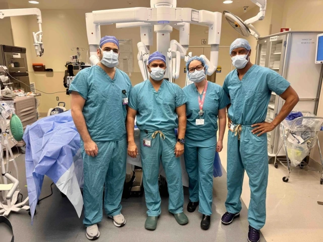 Four healthcare professionals in surgical attire stand in an operating room. They wear blue scrubs, masks, and caps. Behind them is a large piece of medical equipment. The room is well-equipped with medical supplies and monitors.
