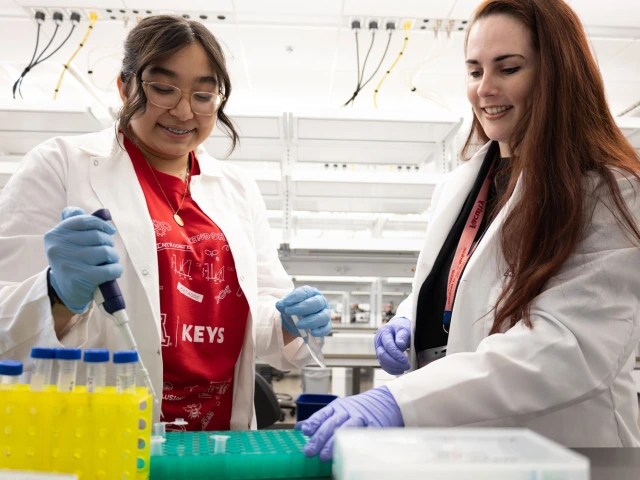 Two individuals in lab coats are working in a laboratory setting. One person is using a pipette with yellow liquid in vials on the table. 