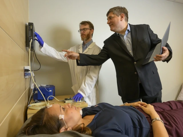 A person is lying on a medical examination table with electrodes attached to their forehead. Two individuals, one in a lab coat using a device and the other in a suit holding a laptop, are next to the table. 