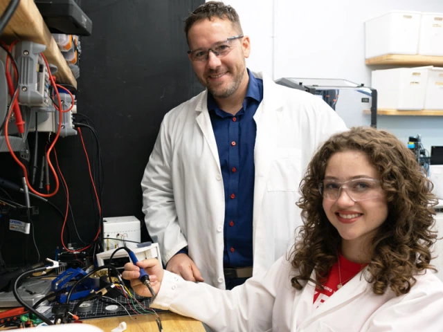Two people in lab coats are in a laboratory setting, surrounded by electronic equipment and wires.