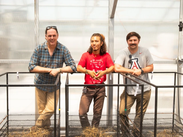 Three individuals stand inside a greenhouse, leaning on metal railings. 