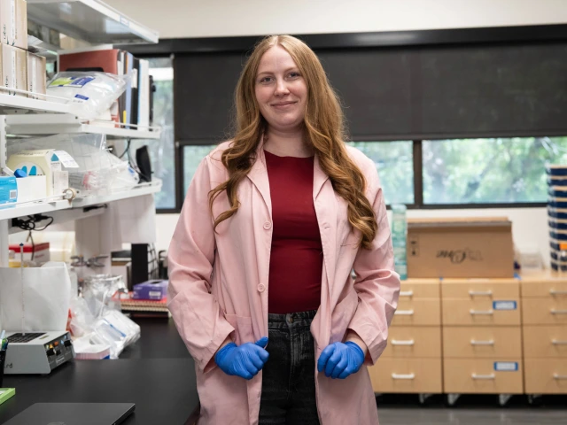 A person stands confidently in a laboratory setting, wearing a pink lab coat and blue gloves. The laboratory is filled with equipment, shelves, and supplies.
