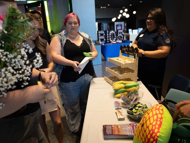 A group of people stands around a table at a convention, where various items like corn-shaped stress toys and brochures are displayed.