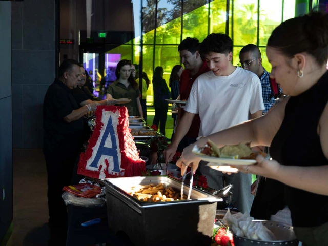 A group of people standing in a buffet line, serving themselves food from trays. There is a cake with a large letter "A" displayed prominently.