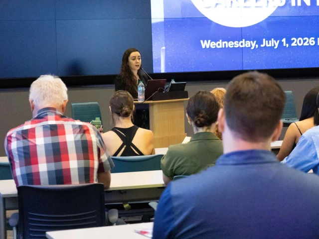 A person stands at a podium giving a presentation in a conference room, with an audience seated at tables. 