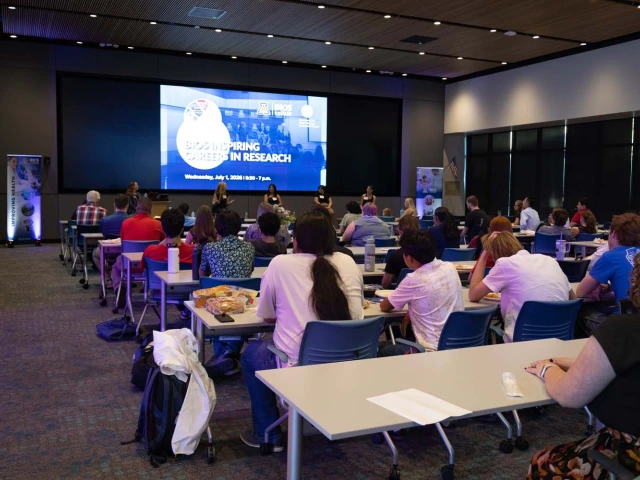 A conference room filled with attendees seated at tables, facing a panel on stage. 