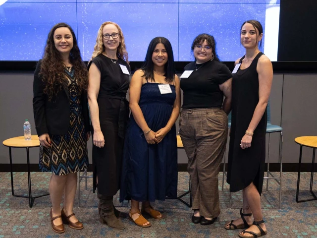 A group of five people is standing together indoors, smiling at the camera.