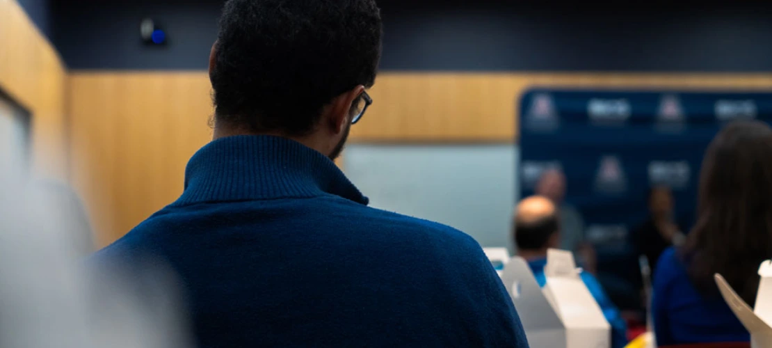 Person with curly hair sitting in a conference room, viewed from behind, focusing on a presentation at the front.