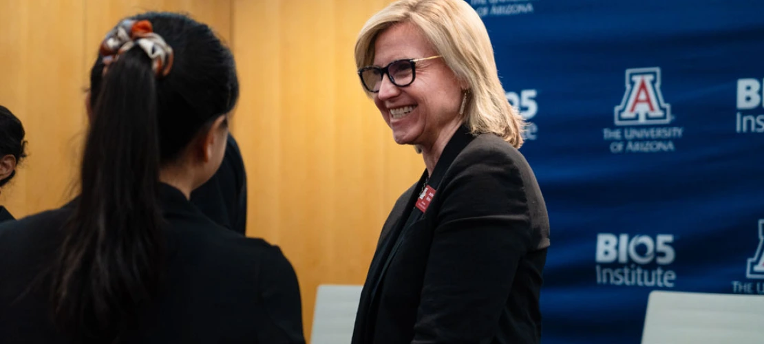 Two individuals engaging in conversation at a University of Arizona BIO5 Institute event. One person, seen from the back, has shoulder-length hair tied with a patterned scarf, while the other, facing forward, is smiling and has short blonde hair, wearing glasses and a name tag. The background features banners with the University of Arizona and BIO5 Institute logos.