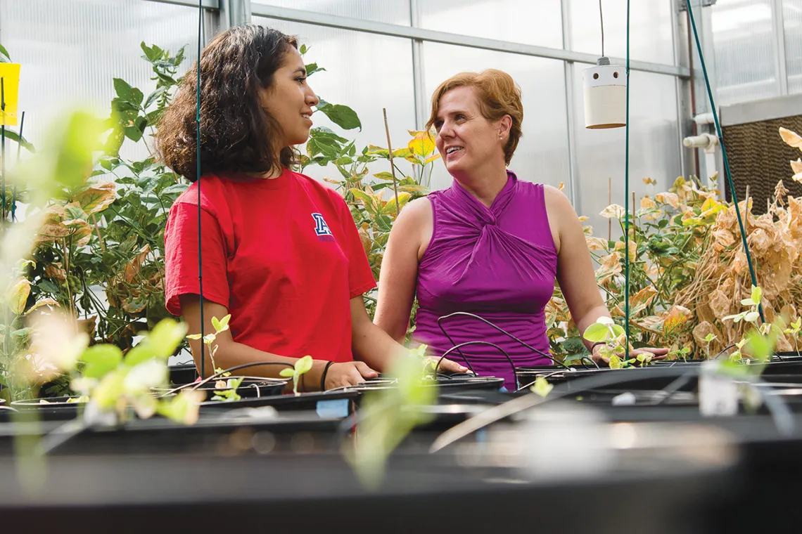 A researcher in a greenhouse with a student next to pots with plants 