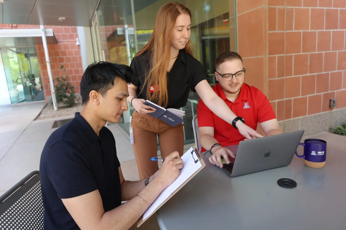 Three students, one standing and two sitting at a table outside a building, are working on a project together with laptops and notebooks.