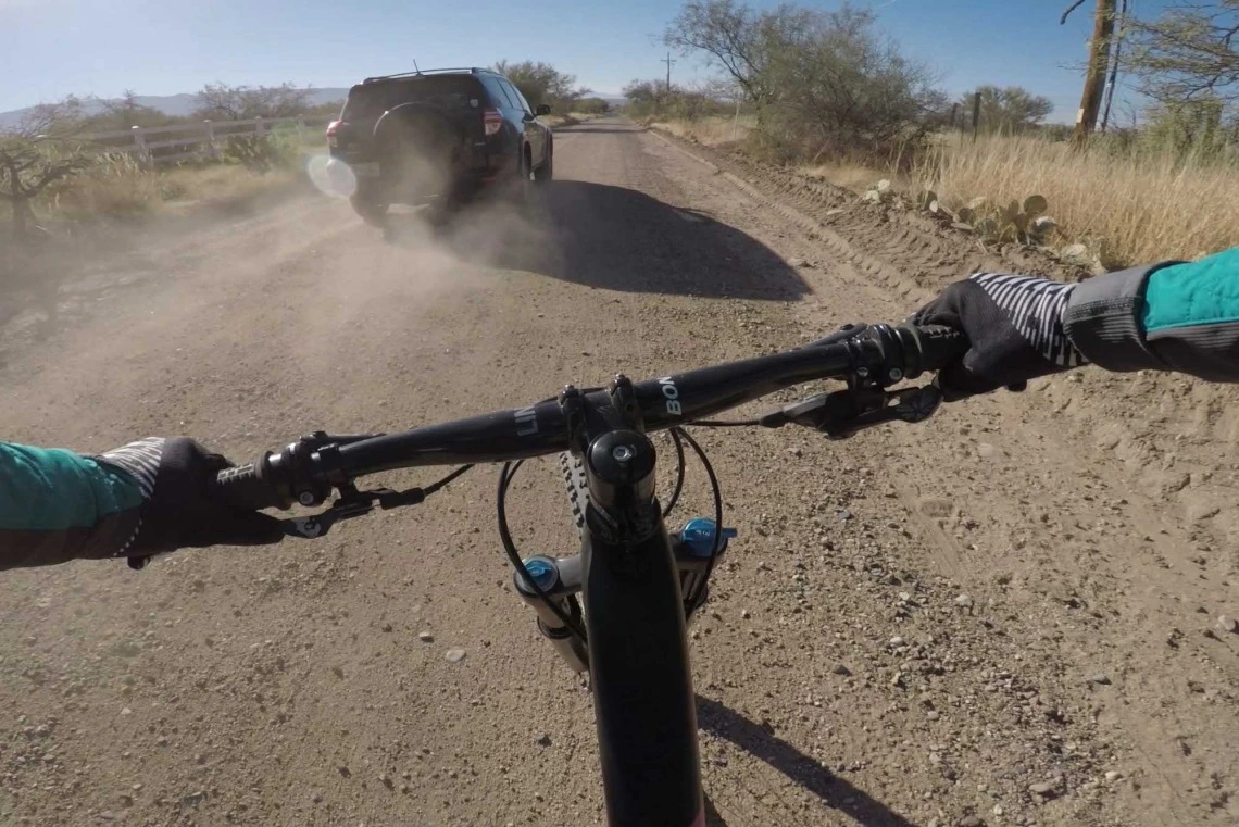 First-person view from a cyclist riding on a dusty road behind a moving vehicle, with handlebars and the bike’s front wheel visible in the forefront.