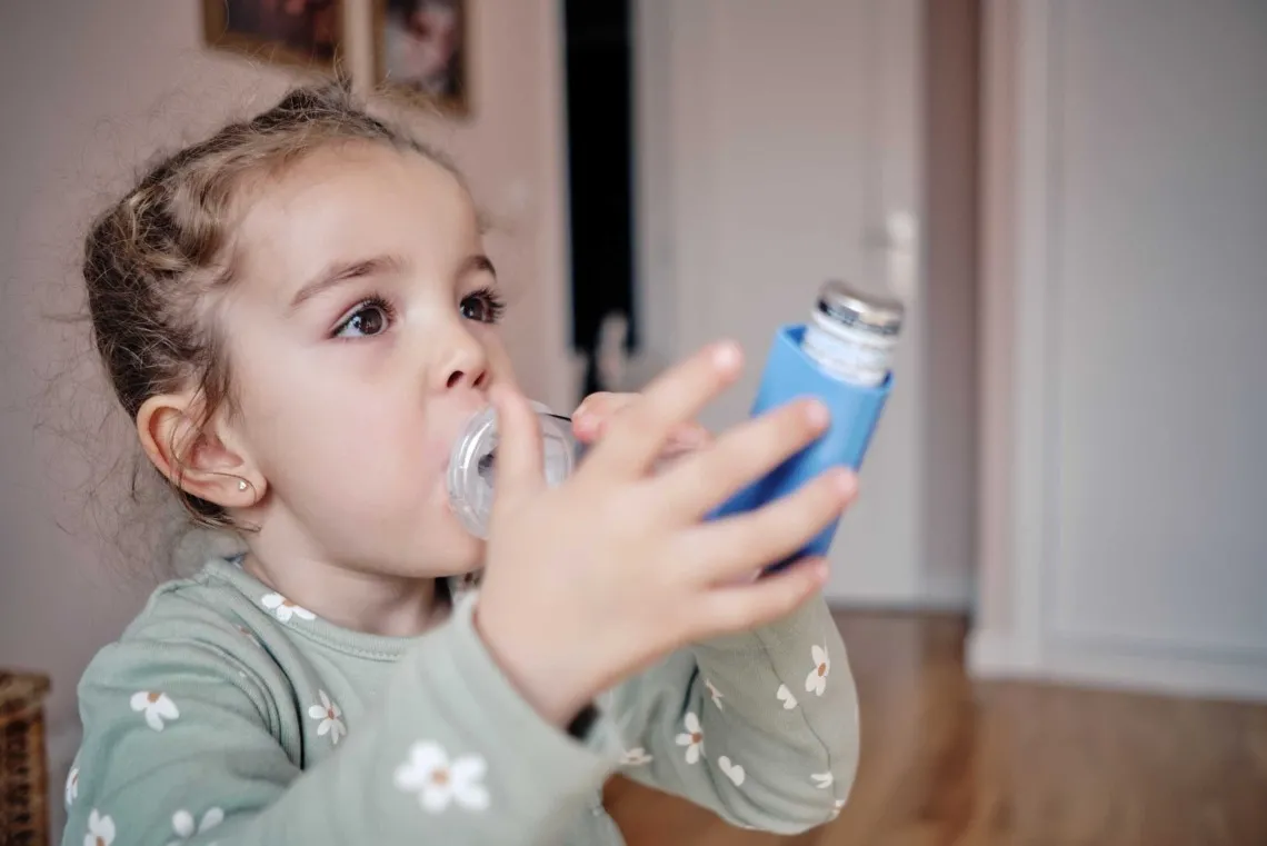 A young child drinks water from a blue insulated bottle. The child has pigtails and is wearing a light-colored shirt with flower patterns. The background shows a softly focused home interior.