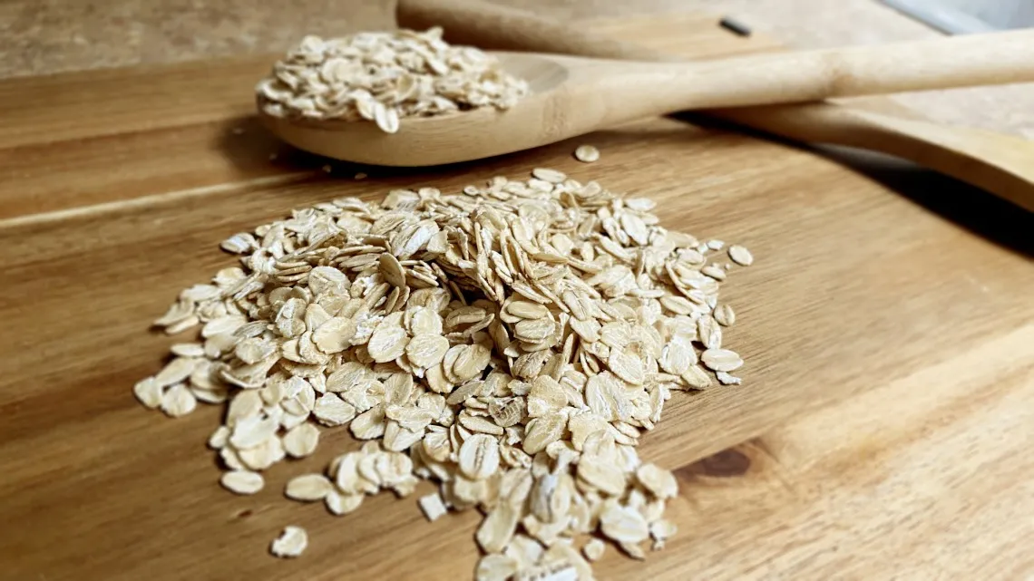 A close-up image of dry rolled oats scattered on a wooden surface, with a wooden spoon also containing oats resting on the surface. The focus is on the texture and natural appearance of the oats.